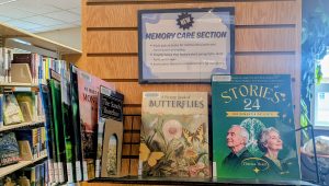 The image showcases a section of a library dedicated to memory care. At the top of the display, there is a sign that reads "NEW MEMORY CARE SECTION," highlighting resources designed to support individuals dealing with dementia and Alzheimer’s. Below the sign, bullet points outline the types of materials available, including adult picture books for mental stimulation and conversation prompting, chapter books with short paragraphs and large fonts, and caregiver resources.

In the foreground, three books are prominently featured. The first is titled “A Picture Book of BUTTERFLIES,” illustrated with colorful butterflies against a floral backdrop. To its right is a green book titled “STORIES 24 FOR DEMENTIA PATIENTS” by Theresa Heart, which features an illustration of an older couple with a brain motif behind them. Several other books in various colors and sizes are partially visible in the background on shelves, indicating a well-stocked library.
Transcribed Text:

NEW MEMORY CARE SECTION

Adult picture books for mental stimulation and conversation prompting
Chapter books that feature short paragraphs, large fonts and images
Books about dementia and Alzheimer's for caregivers